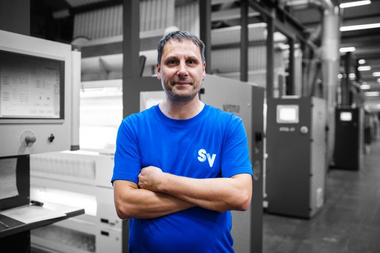 Mitarbeiter ralf steht in blauemn Spandauer velours T-Shirt und verschränkten Armen nett lächelnd vor einer Spannrahmenmaschine ,der Hintergrund ist schawrz-weiß.