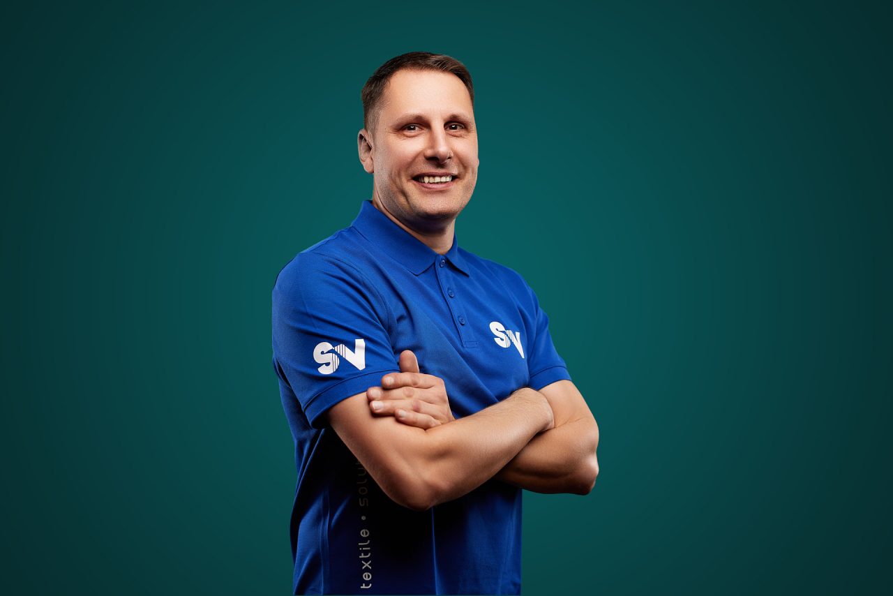 Mitarbeiter Ralf Matthes von Spandauer Velours lächelt mit verschränkten Armen vor einem grünen Hintergrund in die Kamera. Er trägt ein blaues Shirt mit der weiße Aufschrift "SV"