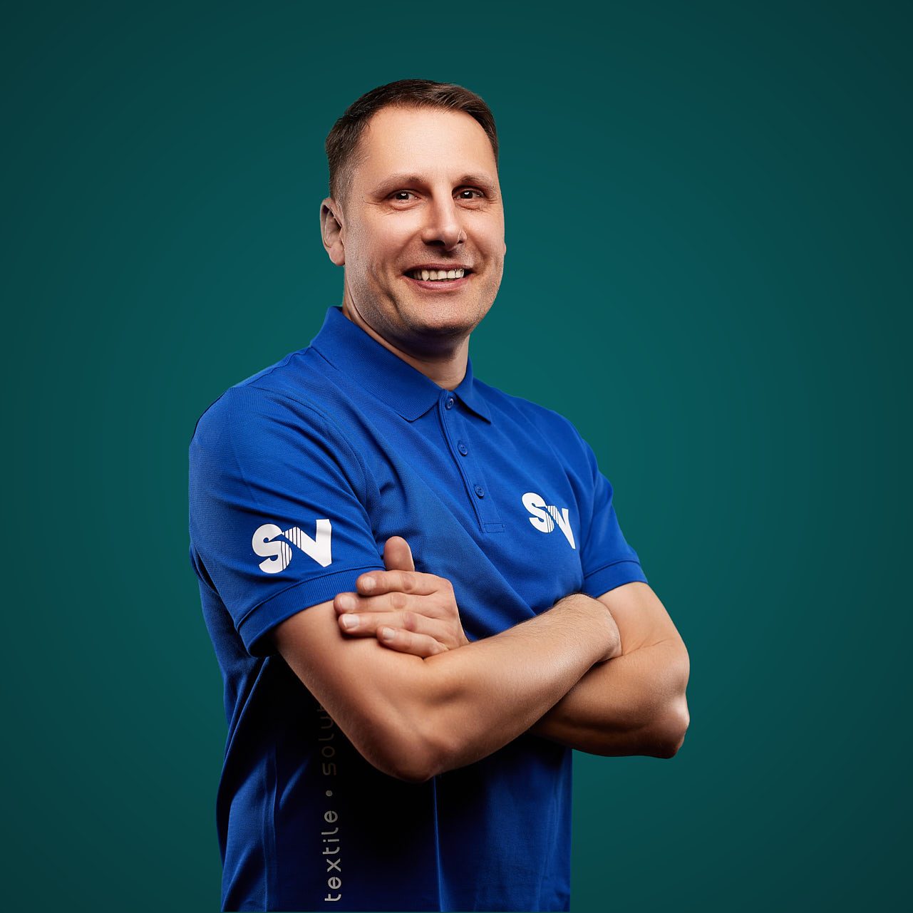 Mitarbeiter Ralf Matthes von Spandauer Velours lächelt mit verschränkten Armen vor einem grünen Hintergrund in die Kamera. Er trägt ein blaues Shirt mit der weiße Aufschrift "SV"