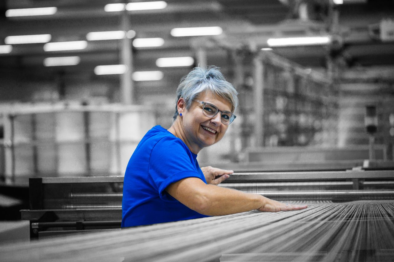 Mitarbeiterin von Spandauer Velours steht in blauem Shirt in der Produktion an einer Stoffmaschine, Hintergund ist schwarz-weiß