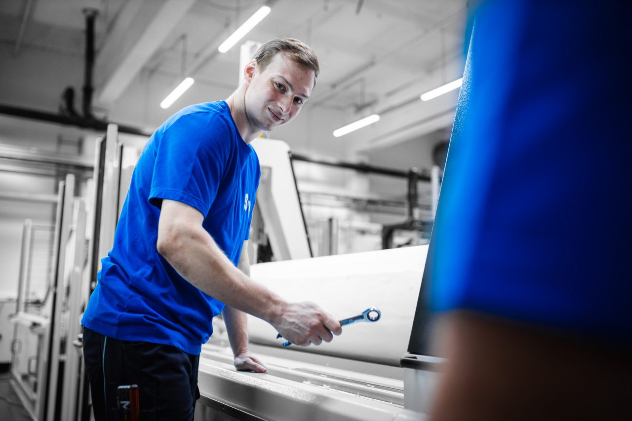 Ein junger Mann in einem blauen T-Shirt arbeitet in einer modern Industriehalle bei Spandauer Verlours. Er blickt lächelnd in die Kamera, während er einen Schraubenschlüssel in der Hand hält.
