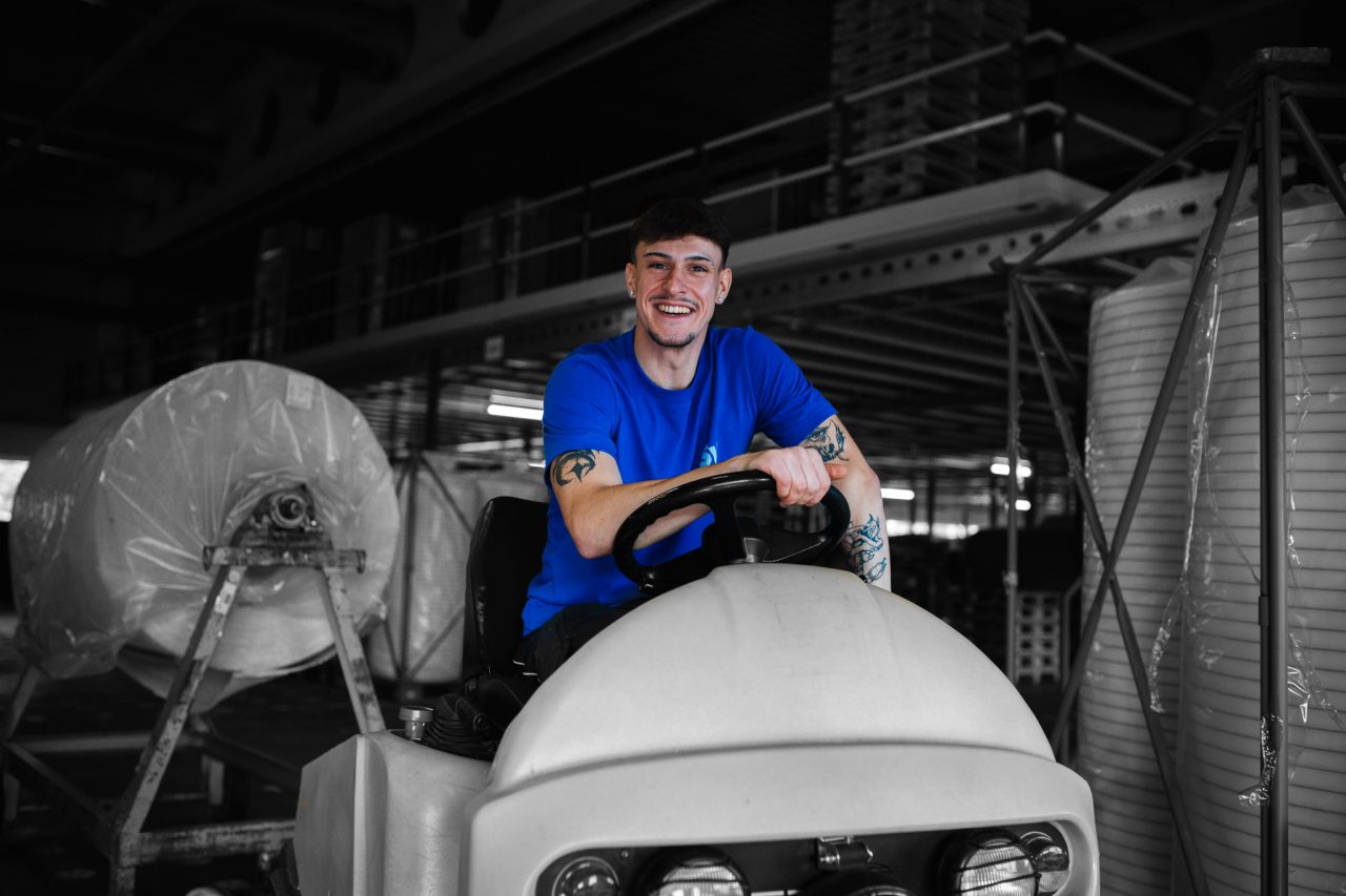 Mitarbeiter Max auf einem Transportmittel n der Verpackungsabteilung lächelt in blauem Spandauer Velours Shirt in die Kamera.